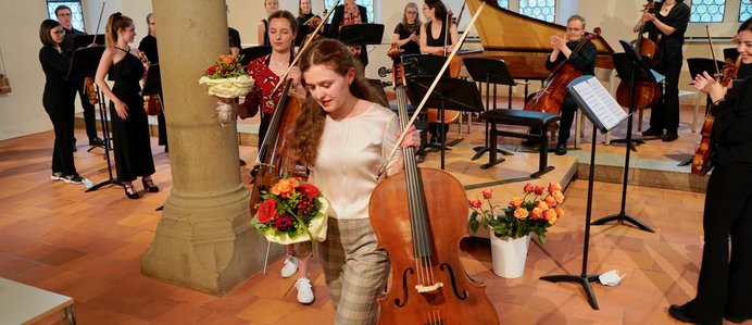 Viele Personen mit Streichinstrumenten am Stehen nach einem Konzert. Zwei Personen laufen mit Blume und Instrument in der Hand davon.
