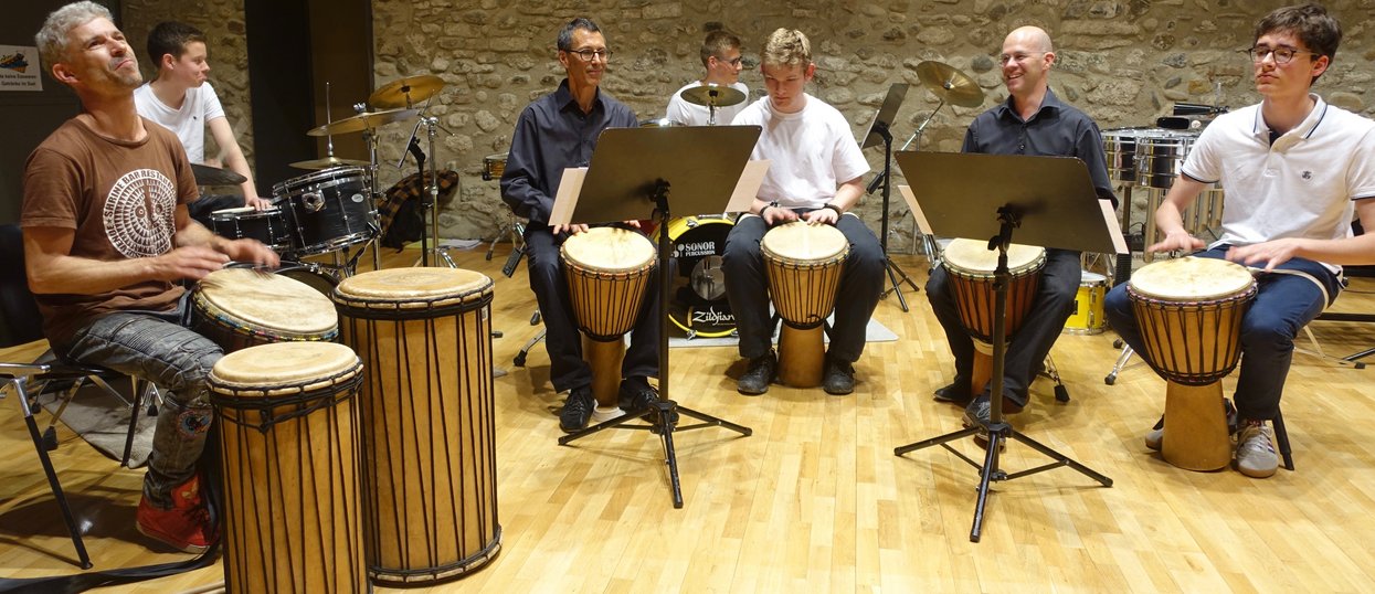 Mehrere Personen in einem Halbkreis spielen gemeinsam auf Trommeln Musik. Im Hintergrund spielen zwei Personen auf dem Schlagzeug.